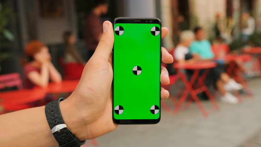 Man holding black smartphone with green screen on street cafe background. Close up. Chroma key. Tracking motion.