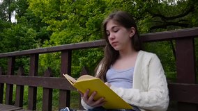 Portrait of serious teenage girl reading book and turning page leaning sitting on the bench in forest in spring, studying outdoor. HD video footage shooting of static camera.