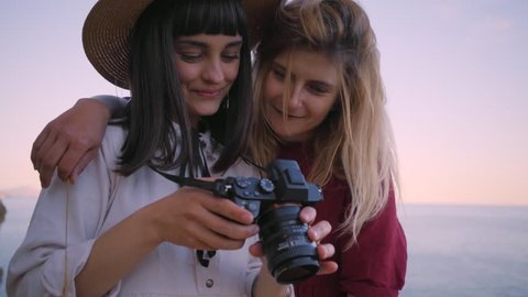 Portrait of two best friends girls, brunette and blonde, smile and laugh, looking photos each of other on their small digital camera with vintage lens, taken at summer evening on sea background