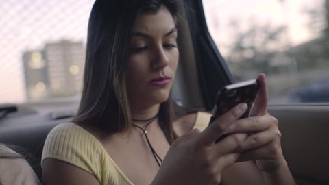 Friends Use A Rideshare, Closeup Of Hispanic Young Woman Using Her Smartphone In Back Seat Of Moving Car