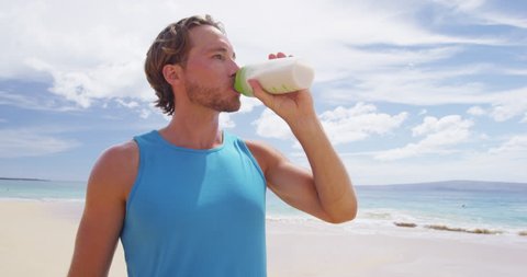 Man drinking protein drink after shaking sports bottle with protein powder mix outside on beach.