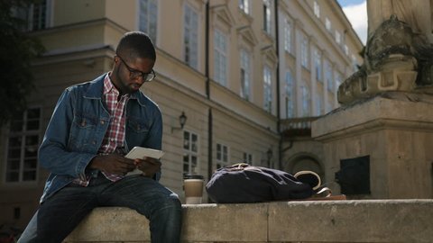 African man in glasses using tablet for chatting with friends on city background and drinking coffee.