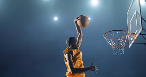 Close up image of professional basketball player making slam dunk during basketball game in floodlight basketball court. The player is wearing unbranded sport clothes.