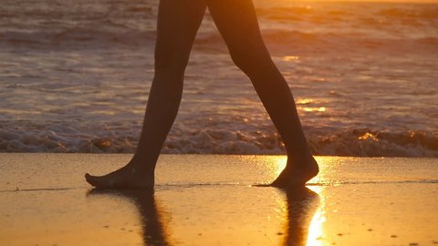 Female feet of hiker tourist walking barefoot on shore at sunset. Legs of young woman going along ocean beach during sunrise. Girl stepping on wet sand of shoreline. Slow motion Close up