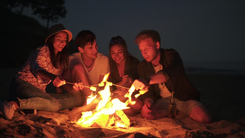 Young cheerful friends sitting by the fire on the beach in the evening, cooking marshmallow on sticks together. Shot in 4k Royalty-Free Stock Footage #28204012