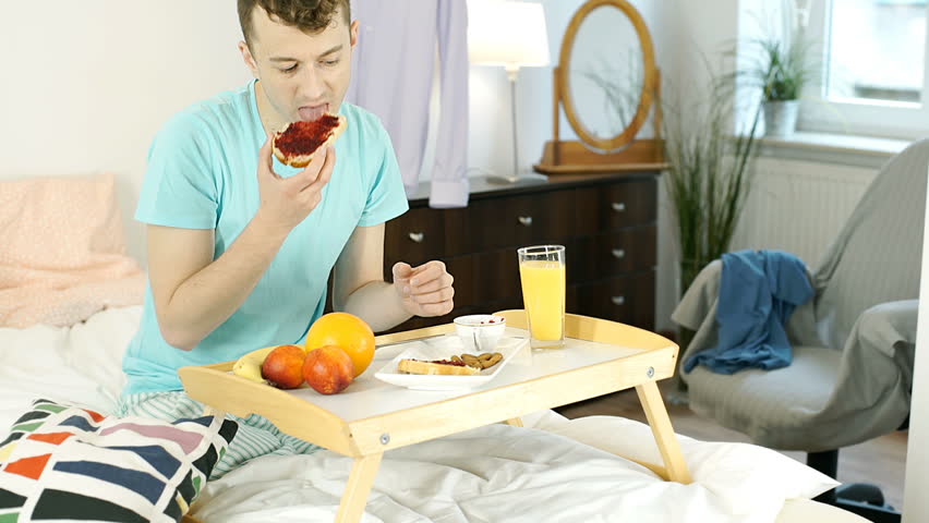 Young man eating breakfast in bed and smiling to the camera, steadycam shot
