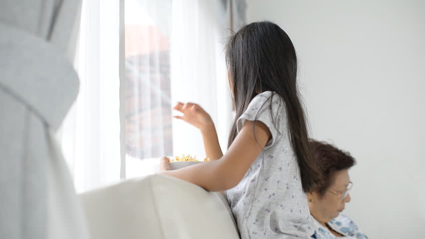 Happy Asian girl eating popcorn and looking out window in raning day
