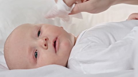 Closeup of lovely little baby lying on bed while mom wiping his face with napkin