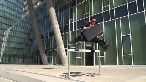 slow motion hurdler in business suit outdoors at office buildings running over hurdle with briefcase
