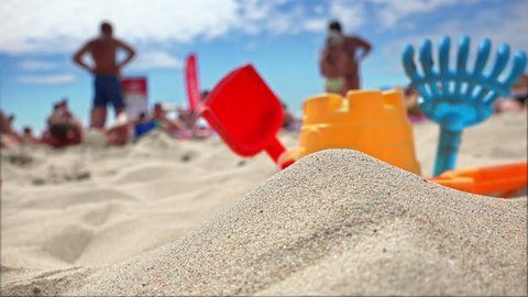 Children's beach toys - buckets, spade and shovel on sand on a sunny day with tourists blurred at background