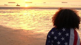 4K video clip of mixed race African American girl teenager female young woman holding an American US Stars and Stripes flag on a beach at sunset or sunrise