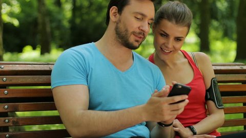 Happy young couple sitting on a bench after morning run and using smart phone. Woman and man watching video, photos.