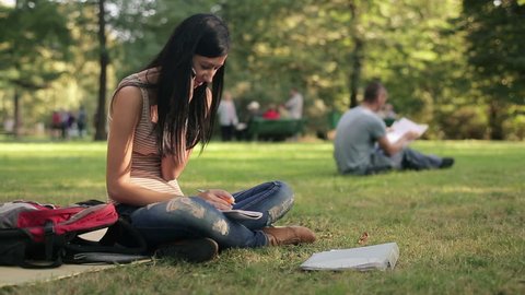 Female student doing homework and talking on cellphone in the park
