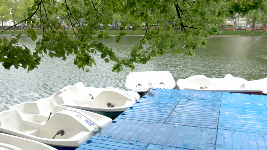 A station for renting boats on a city pond. A fine rain comes on a summer day. Drops of rain fall into a city pond, on boats. Beautiful background for text.