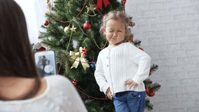 Cheerful little girl dancing and playing the ape near Christmas tree while mother shooting her photo and video by smart phone. Funny video in 4k slow motion.