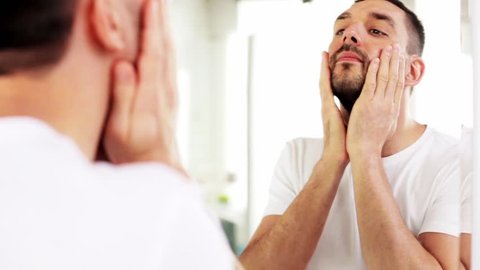 beauty, skin care and people concept - smiling young man applying after shave oil to face and looking to mirror at home bathroom