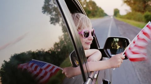 A blond little girl in pink glasses looks out the window of the car. In his hand holds the American flag