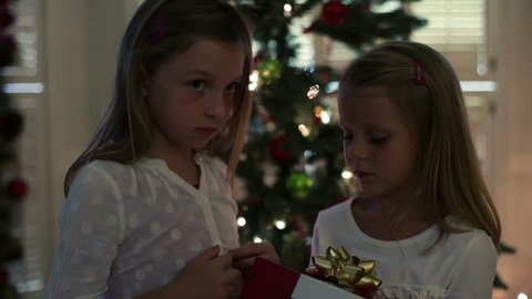 Two cute little blond girls standing in front of a Christmas tree open a box that reveals a bright light from inside.