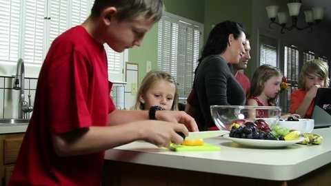 Big brother prepares healthy snack for little sister while rest of family looks at something on laptop.