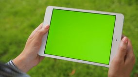 Female hands holding white tablet computer device with green screen on green grass background. Outdoors in park. Woman scrolling, zooming, tapping on touchscreen. Chroma key. Close up.