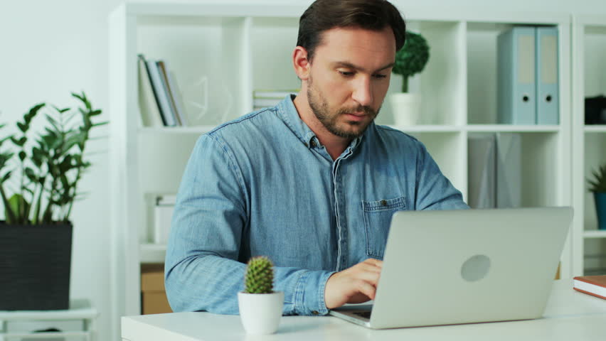 Tired young business man in blue shirt finishing work on the laptop in the office, having annoying back pain.
