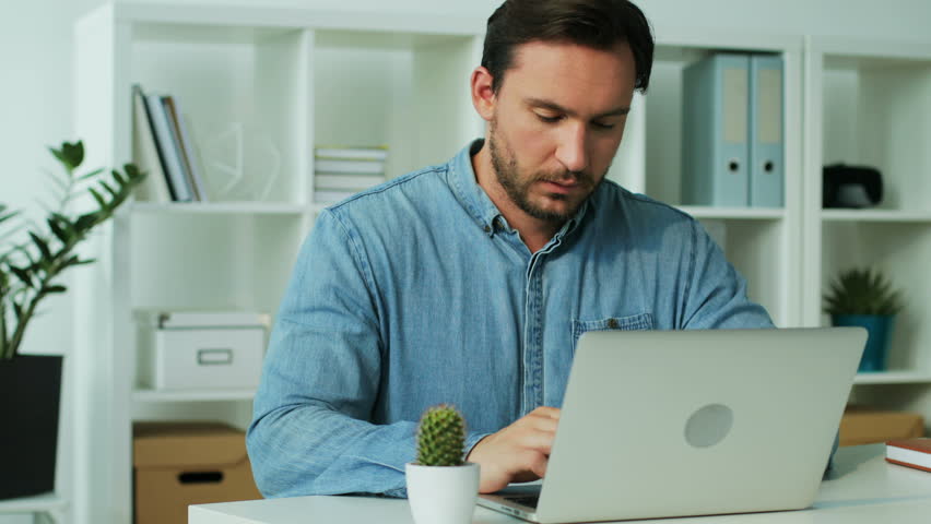 Serious young business man in blue shirt finishing work on the laptop in the office, having annoying headache.