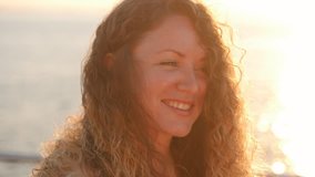 Portrait of young attractive woman with curly red hair standing near sea or ocean in sunrise or sunset light. The girl looks at the camera, then straightens her hair and looks at the sun. Slow motion.