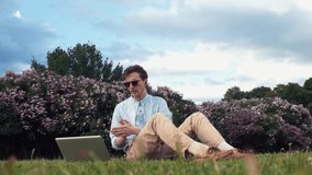 Caucasian young man in sunglasses having video chat at laptop sitting on green grass at park. Summer day. Freelancer working outdoors. Blue sky and bright sun on the background.