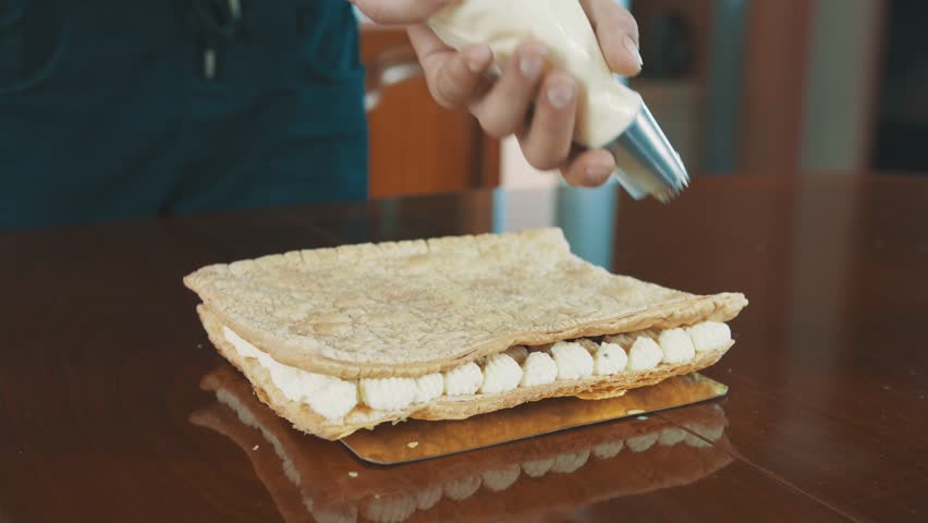 Confectioner young buffed caucasian bearded man squeezes whiped creme puffs in line out sleeve onto cake layer on kitchen table