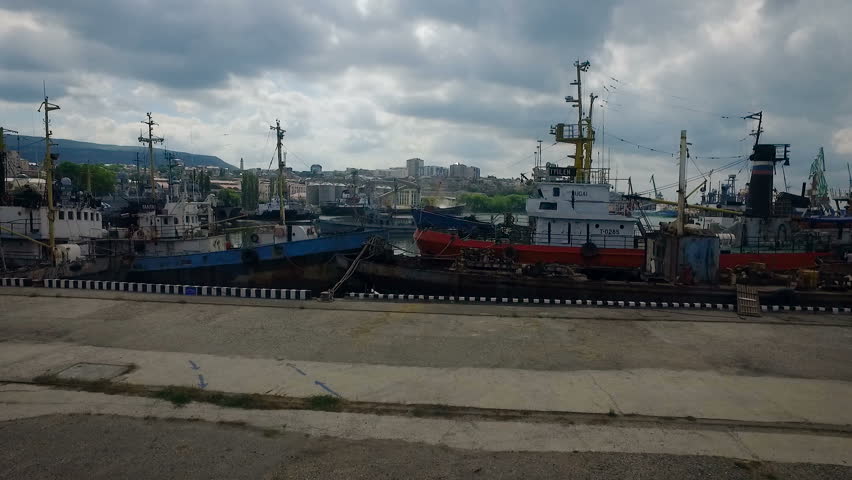 Sea Port on Bay. Town on Green Hill. Cargo Boats Staying in Dock. Vessels at Pier. Cistern on Shore. Cloudy Summer Day