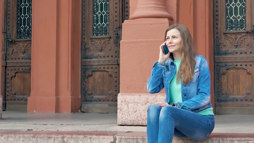 Pretty girl sits on the steps in a cotton jacket and talking on smartphone. Vintage building in the background.