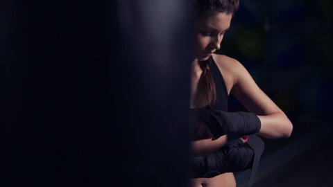 Young beautiful woman wrapping hands with black boxing wraps in dark room. Slowmotion shot