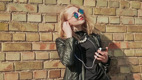Stylish young blonde in sunglasses and leather jacket listening to music on bluetooth headphones in a mobile phone. Enjoy music. Spring. Background of a brick wall.