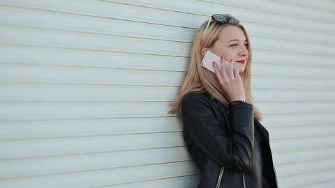 Beautiful young blonde girl in leather jacket and sunglasses talking on mobile phone outdoors. Against a background of white horizontal roller blinds.