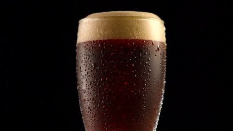 Dark Beer closeup. Pint of cold Craft beer isolated on matte black background, rotation 360 degrees. Glass of beer with water drops. 4K UHD video 3840x2160