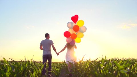 Beauty Couple with colorful air balloons run away on summer field, holding hands and smiling. Happy family enjoying nature together, healthy lifestyle concept. Girlfriend, boyfriend. Slow motion 4K