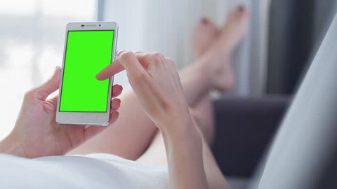 Young Woman in white top laying on couch uses mobile phone with pre-keyed green screen. Few types of gestures - scrolling up and down, tapping, zoom in and out. Perfect for screen compositing