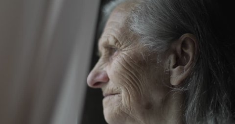 Close-up face of sad old woman with deep wrinkles. Grandmother with gray hair looks out the window and smile. 4K footage. 