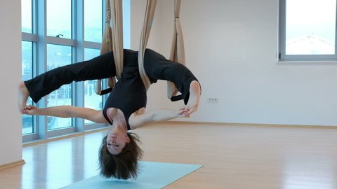Young woman hanging upside down in hammock for yoga indoors. She slowly swings body with her legs wide apart and holds them with her fingers then she fully straightens and leans her hands on floor