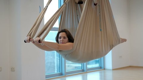 Young brunette hanging in hammock for yoga indoors. Attractive woman lying down on her stomach in special canvas and holding hands with hinges easily swings and hovers over floor. She is training in