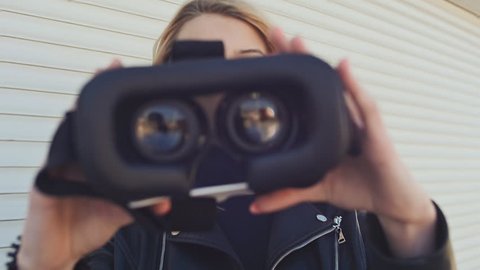 A young stylish blonde in a leather jacket near horizontal roller blinds brings virtual glasses to the camera.