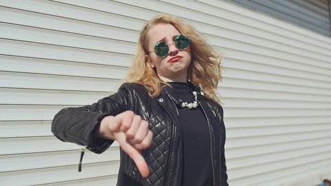 Stylish young girl in a leather jacket and sunglasses shows first the gesture of the finger down, later the gesture of the thumbs up. Background of white horizontal rolling shutters.
