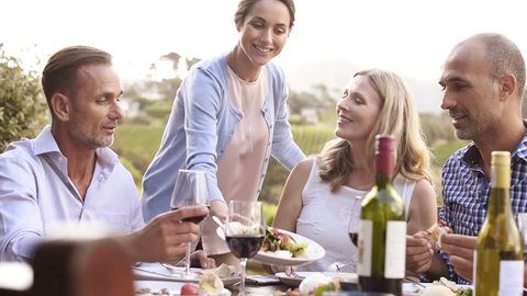 Happy smiling woman serving salad to friends and family outdoor in a sunset. Happy mature friends having dinner together. Senior men and middle aged women having fun while eating together at vineyard.