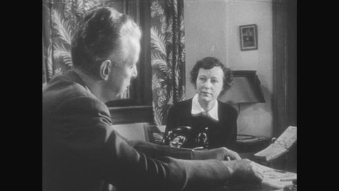 1950s: Man in office, seated at desk, talks to woman who is seated across desk, referencing papers on the desk. Brief glimpse of another woman not in the office.
