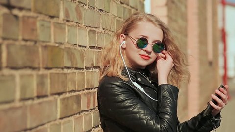 Stylish young blonde in sunglasses and leather jacket listening to music on headphones in a mobile phone. Enjoy music. Background of a brick wall.