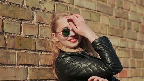 Stylish young blonde in sunglasses and black leather jacket near a brick wall on the street.