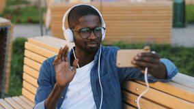 Attractive african man in glasses with earphones using smart phone for video chatting with friend siting on the bench in city.