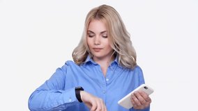 Young serious woman using and tuning smartwatches and mobile phone isolated over white