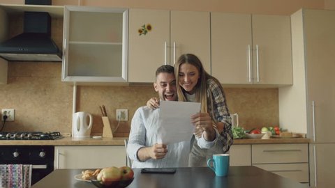 Attractive happy couple recieve good news unfolding letter in the kitchen while have breakfast at home