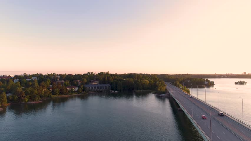 Aerial warm yellow scene drone flying towards above sea, bridge with driving cars beside on a summer evening around sunset. Lauttasaari, Länsiväylä, Lapinlahti bridge, Helsinki, Finland, Scandinavia.